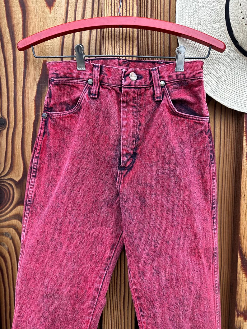 May include: A pair of acid-wash pink denim jeans hanging on a red hanger. The jeans have a classic five-pocket design with a button and zipper closure. The denim has a faded, distressed look.