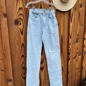 May include: Pale blue denim jeans hanging on a clear hanger. The jeans have a high waist and a straight leg. A straw hat with a black band hangs on the wooden wall behind the jeans.