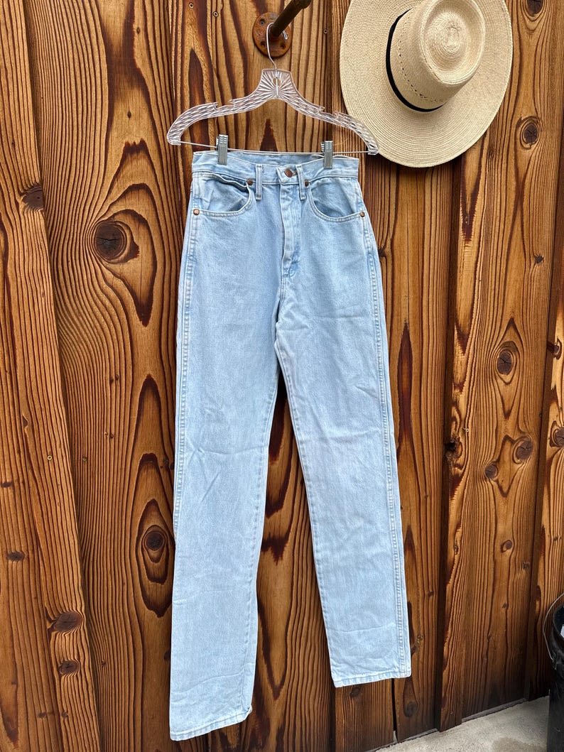 May include: Pale blue denim jeans hanging on a clear hanger. The jeans have a high waist and a straight leg. A straw hat with a black band hangs on the wooden wall behind the jeans.