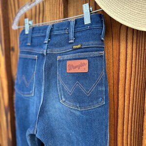 Puede incluir: Un par de pantalones vaqueros de mezclilla azul oscuro con un parche de cuero marr&oacute;n Wrangler en el bolsillo trasero.