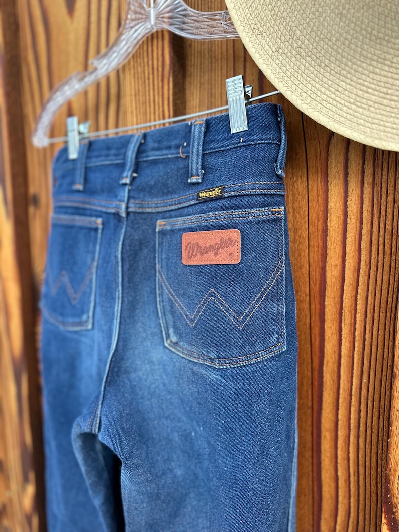 Puede incluir: Un par de pantalones vaqueros de mezclilla azul oscuro con un parche de cuero marr&oacute;n Wrangler en el bolsillo trasero.
