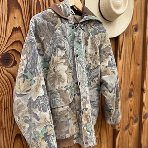 May include: A camouflage jacket with a hood and pockets hangs on a wooden hanger. The jacket features a pattern of green, brown, and tan leaves and branches. A straw hat with a black band is visible in the background.