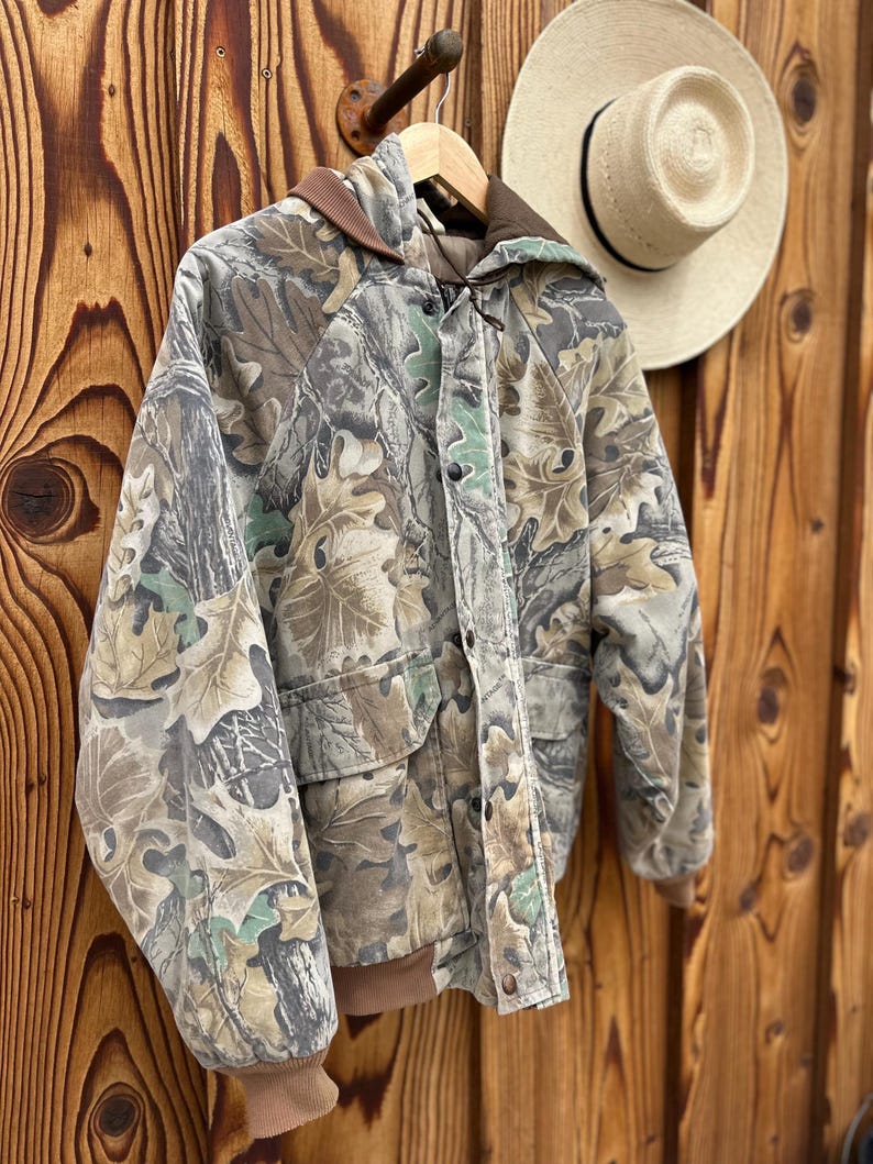 May include: A camouflage jacket with a hood and pockets hangs on a wooden hanger. The jacket features a pattern of green, brown, and tan leaves and branches. A straw hat with a black band is visible in the background.