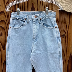 May include: Pale blue denim jeans with a traditional five-pocket layout, suspended on a clear plastic hanger. The jeans feature a high waist and a straight leg, with copper rivets and a button fastening. The denim has a light wash appearance.