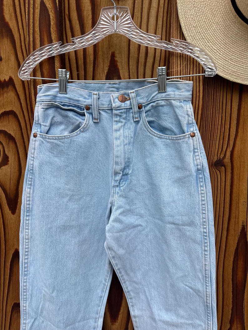 May include: Pale blue denim jeans with a traditional five-pocket layout, suspended on a clear plastic hanger. The jeans feature a high waist and a straight leg, with copper rivets and a button fastening. The denim has a light wash appearance.
