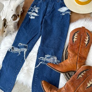 K&ouml;nnte beinhalten: Eine blaue Jeans mit Destroyed-Details, die mehrere Risse und L&ouml;cher im Stoff aufweist. Die Jeans liegt auf einer wei&szlig;en, flauschigen Oberfl&auml;che, zusammen mit einem Paar braunen Leder-Cowboots und einem Strohhut mit gelbem Band.