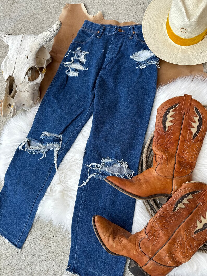 K&ouml;nnte beinhalten: Eine blaue Jeans mit Destroyed-Details, die mehrere Risse und L&ouml;cher im Stoff aufweist. Die Jeans liegt auf einer wei&szlig;en, flauschigen Oberfl&auml;che, zusammen mit einem Paar braunen Leder-Cowboots und einem Strohhut mit gelbem Band.