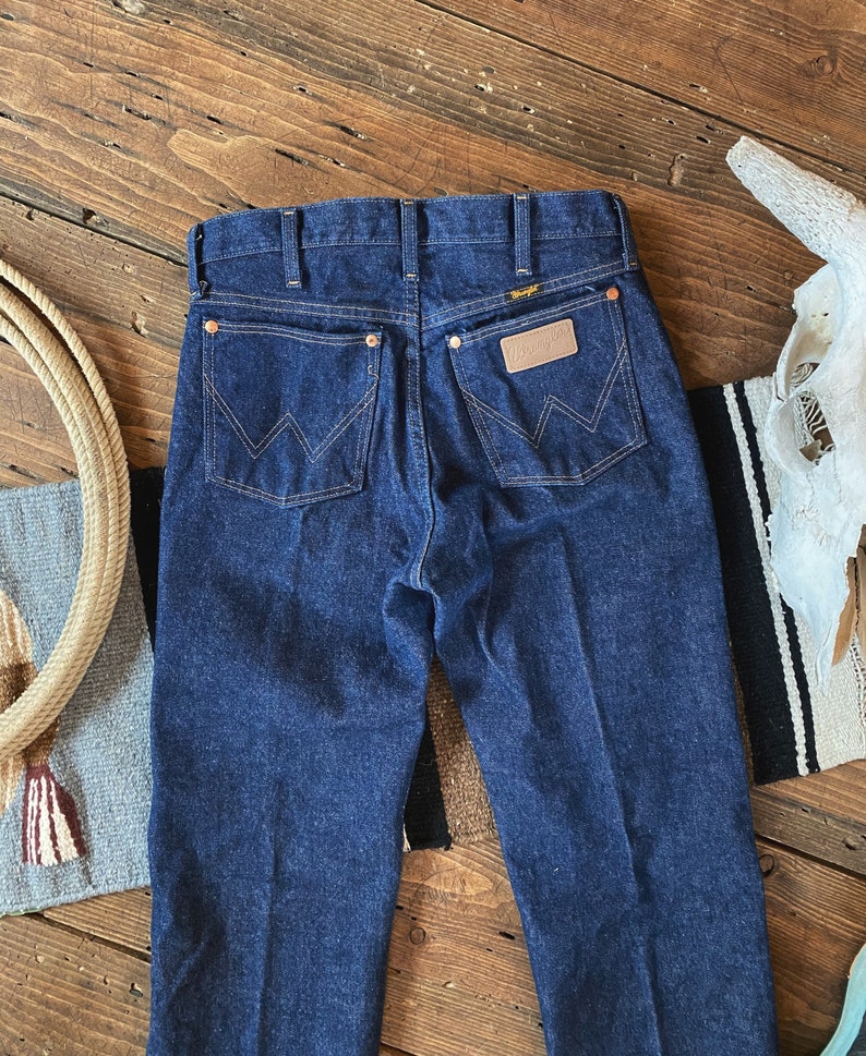 May include: A pair of dark blue denim jeans with a classic straight leg fit. The jeans have a leather patch on the back with the Wrangler logo.