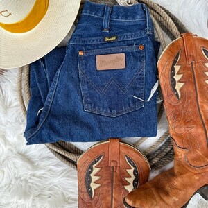 K&ouml;nnte beinhalten: Ein Paar braune Leder-Cowbo Stiefel mit aufwendigen N&auml;hten und wei&szlig;em Hintergrund. Eine blaue Jeans mit einem braunen Leder-Wrangler-Etikett. Ein wei&szlig;er Strohhut mit gelbem Band.