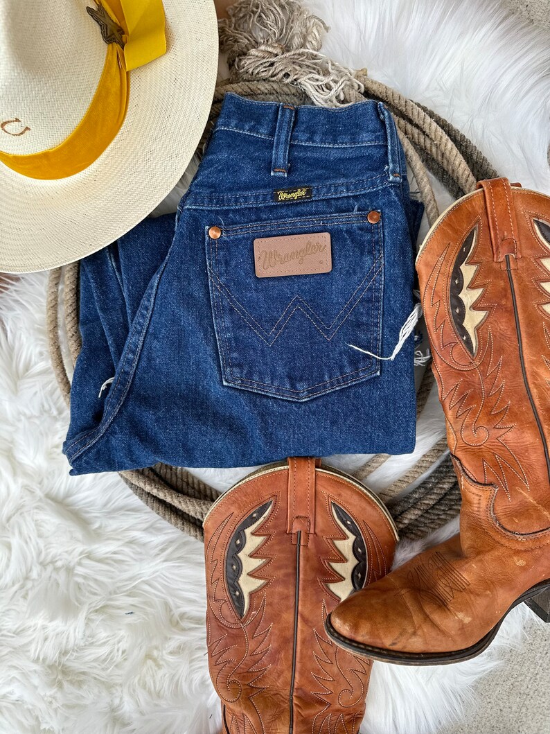 K&ouml;nnte beinhalten: Ein Paar braune Leder-Cowbo Stiefel mit aufwendigen N&auml;hten und wei&szlig;em Hintergrund. Eine blaue Jeans mit einem braunen Leder-Wrangler-Etikett. Ein wei&szlig;er Strohhut mit gelbem Band.