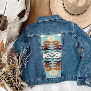 May include: A blue denim jacket with a colorful geometric design on the back. The design features shades of turquoise, brown, and white. The jacket has a classic collar, button closures, and pockets.