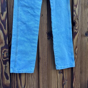 May include: Light blue denim jeans. The jeans have a classic five-pocket design, a straight leg, and a medium wash. The jeans are made of a durable denim fabric and are suitable for everyday wear. The jeans are shown against a wooden background.