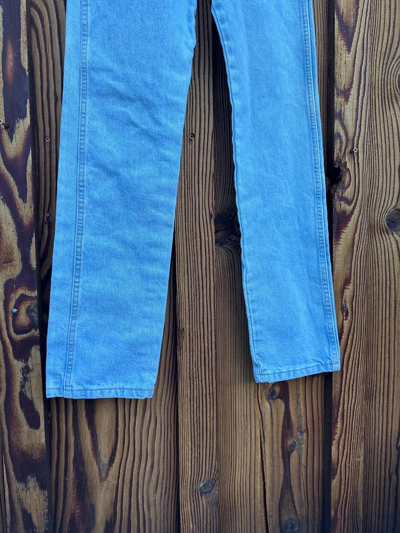 May include: Light blue denim jeans. The jeans have a classic five-pocket design, a straight leg, and a medium wash. The jeans are made of a durable denim fabric and are suitable for everyday wear. The jeans are shown against a wooden background.