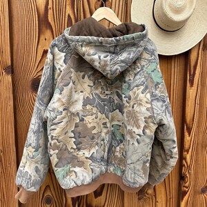 May include: A camouflage jacket with a brown hood and cuffs hangs on a wooden hanger. The jacket features a leaf pattern in shades of green, brown, and gray. A straw hat with a black band rests on the wooden background.