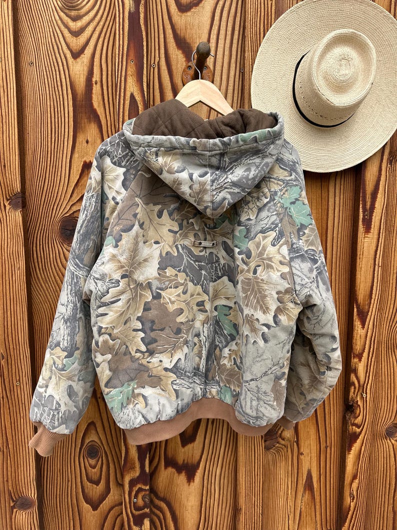 May include: A camouflage jacket with a brown hood and cuffs hangs on a wooden hanger. The jacket features a leaf pattern in shades of green, brown, and gray. A straw hat with a black band rests on the wooden background.