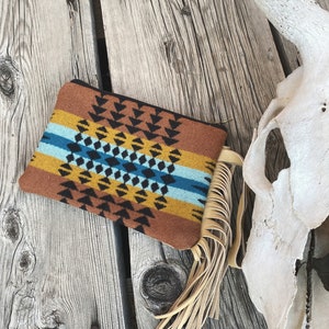 May include: A brown and tan patterned clutch with a fringe tassel. The clutch has a geometric design with black, blue, and yellow accents.