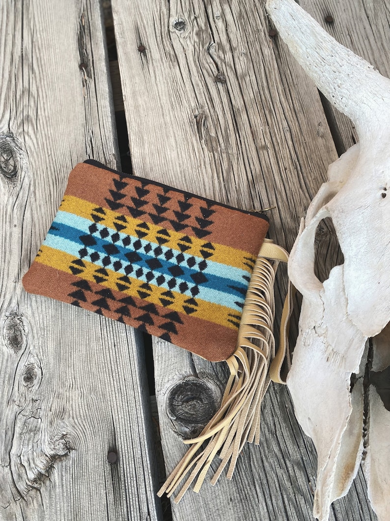 May include: A brown and tan patterned clutch with a fringe tassel. The clutch has a geometric design with black, blue, and yellow accents.