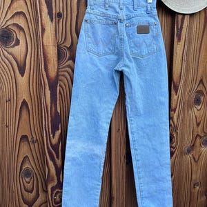 May include: Light blue denim jeans with a classic five-pocket design, featuring the Wrangler logo on the back pockets and a brown leather patch. The jeans are hanging on a hanger against a wooden background.