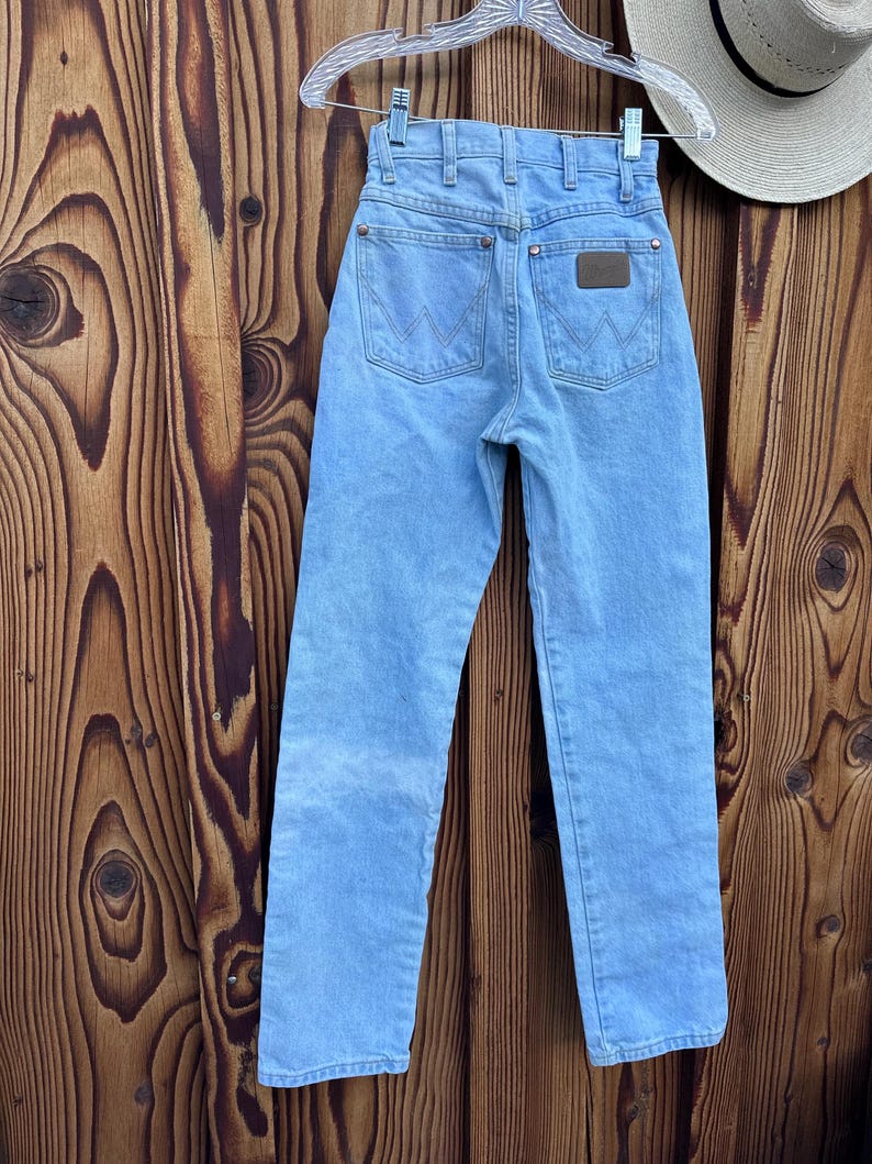 May include: Light blue denim jeans with a classic five-pocket design, featuring the Wrangler logo on the back pockets and a brown leather patch. The jeans are hanging on a hanger against a wooden background.
