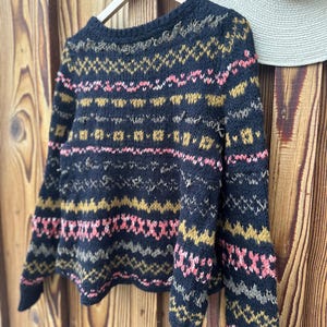 May include: A navy blue sweater with a geometric pattern in pink, gold, and gray. The long-sleeved sweater is hanging on a wooden hanger against a wooden background. A straw hat hangs on the right side of the image.
