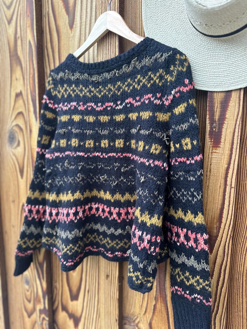 May include: A navy blue sweater with a geometric pattern in pink, gold, and gray. The long-sleeved sweater is hanging on a wooden hanger against a wooden background. A straw hat hangs on the right side of the image.