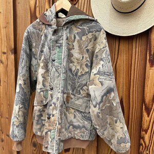 May include: Camouflage print jacket with a brown hood, cuffs, and trim. The jacket has a button-up front, two front pockets, and a hood. A straw hat with a black band hangs nearby. The jacket is hanging on a wooden hanger.