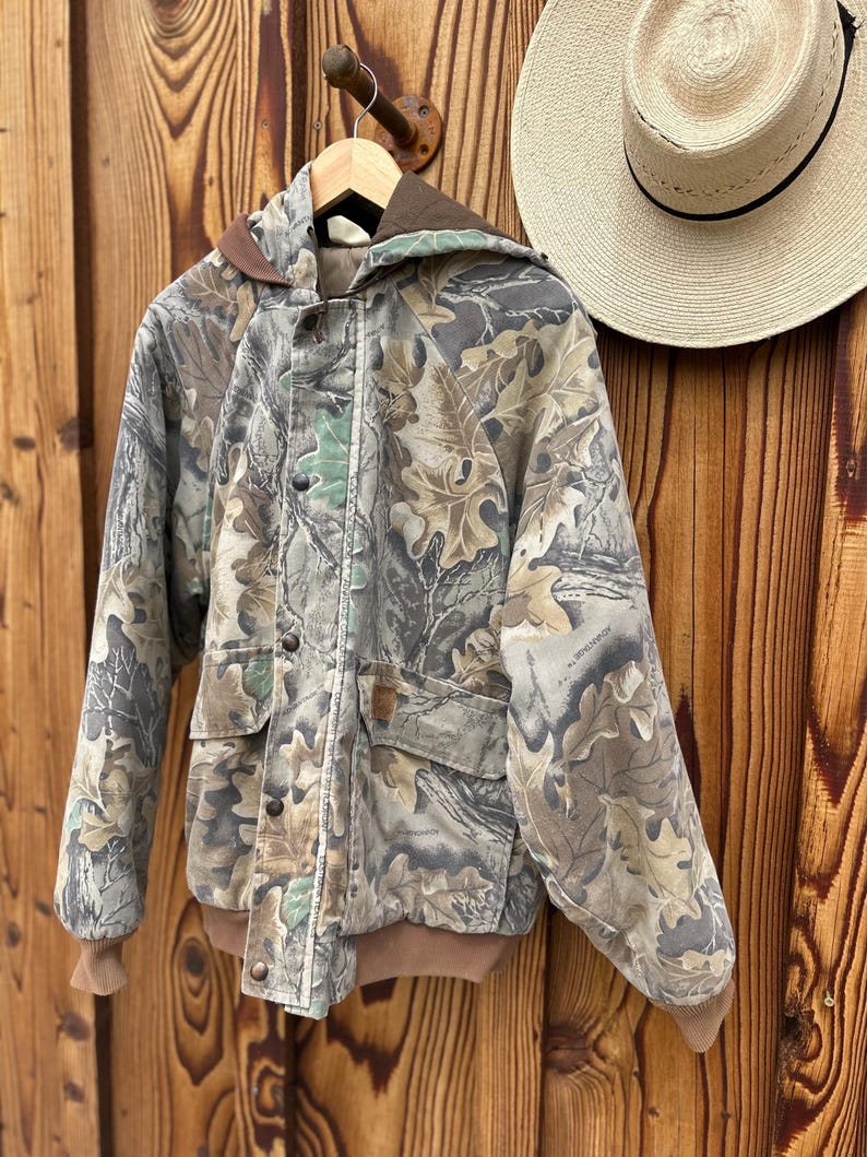 May include: Camouflage print jacket with a brown hood, cuffs, and trim. The jacket has a button-up front, two front pockets, and a hood. A straw hat with a black band hangs nearby. The jacket is hanging on a wooden hanger.