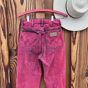 May include: A pair of acid-wash pink Wrangler jeans hangs on a red hanger against a wooden background. The jeans have a classic five-pocket design with the Wrangler logo on the back pocket and a leather patch. A white straw hat is visible in the background.