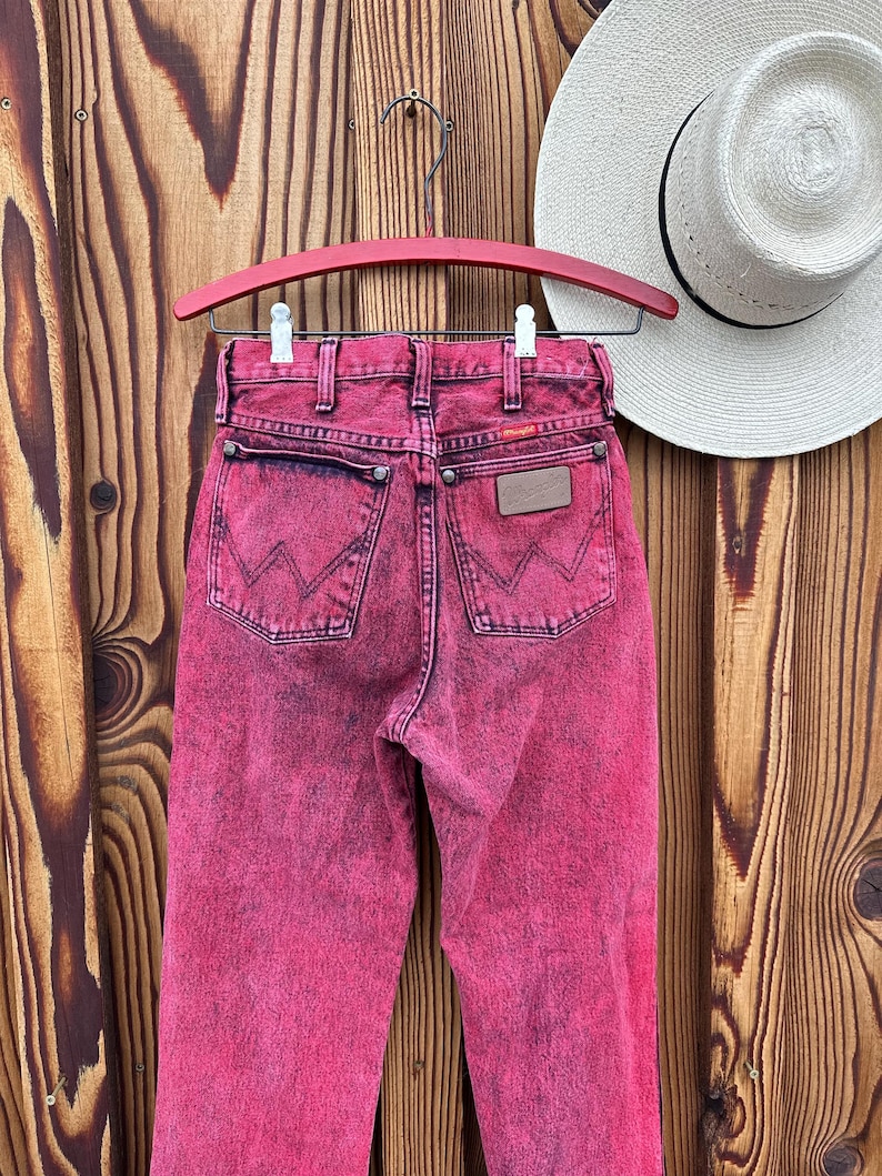 May include: A pair of acid-wash pink Wrangler jeans hangs on a red hanger against a wooden background. The jeans have a classic five-pocket design with the Wrangler logo on the back pocket and a leather patch. A white straw hat is visible in the background.