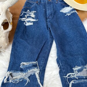 K&ouml;nnte beinhalten: Distressed blaue Jeans aus Denim mit mehreren Rissen und ausgefransten Kanten. Die Jeans hat eine hohe Taille und ein gerades Bein.