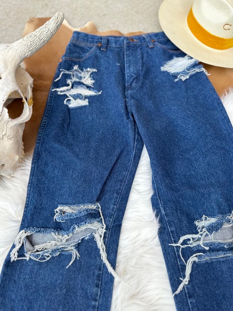 K&ouml;nnte beinhalten: Distressed blaue Jeans aus Denim mit mehreren Rissen und ausgefransten Kanten. Die Jeans hat eine hohe Taille und ein gerades Bein.