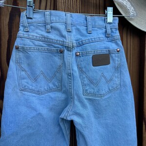 May include: Light blue denim jeans with the Wrangler logo on the back pocket. The jeans have classic five-pocket styling, copper rivets, and a light wash. The jeans are hanging on a clear hanger against a wooden background.