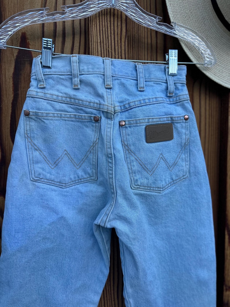 May include: Light blue denim jeans with the Wrangler logo on the back pocket. The jeans have classic five-pocket styling, copper rivets, and a light wash. The jeans are hanging on a clear hanger against a wooden background.