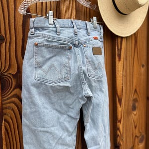 以下が含まれることがあります： ライトブルーのデニムジーンズ。バックポケットにはWranglerのロゴ、茶色の革パッチが付いています。透明なプラスチックハンガーに吊るされ、木製の背景にあります。黒いバンドが付いた麦わら帽子も写っています。