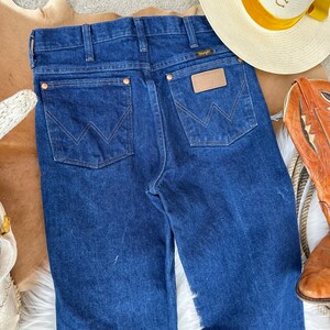 May include: A pair of dark blue denim jeans with a classic five-pocket style. The jeans feature a leather patch with the Wrangler logo on the back right pocket.