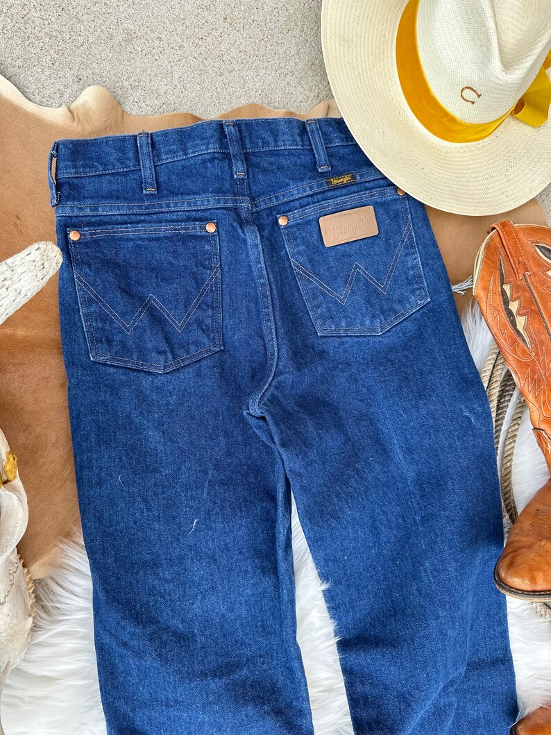 May include: A pair of dark blue denim jeans with a classic five-pocket style. The jeans feature a leather patch with the Wrangler logo on the back right pocket.