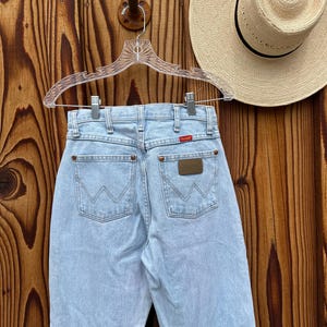 May include: Pale blue denim jeans with the Wrangler logo on the back pockets. The jeans are hanging on a clear hanger against a wooden background. A straw hat with a black band is visible.