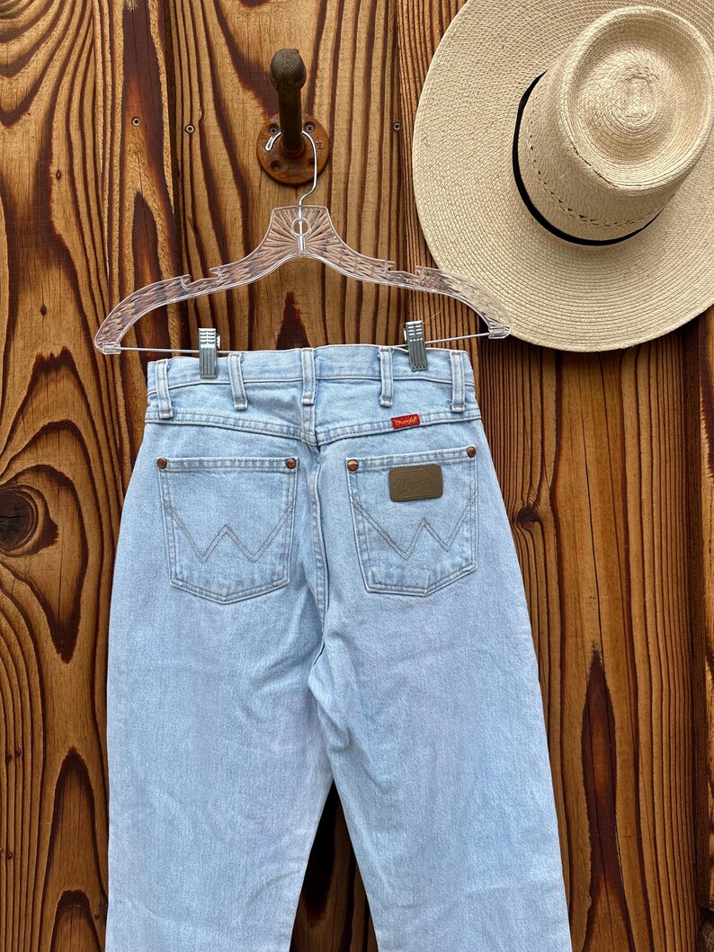 May include: Pale blue denim jeans with the Wrangler logo on the back pockets. The jeans are hanging on a clear hanger against a wooden background. A straw hat with a black band is visible.