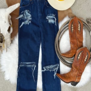 May include: Distressed blue denim jeans with frayed edges and rips at the knees. The jeans are laid flat on a white surface with a cowboy hat and boots in the background.