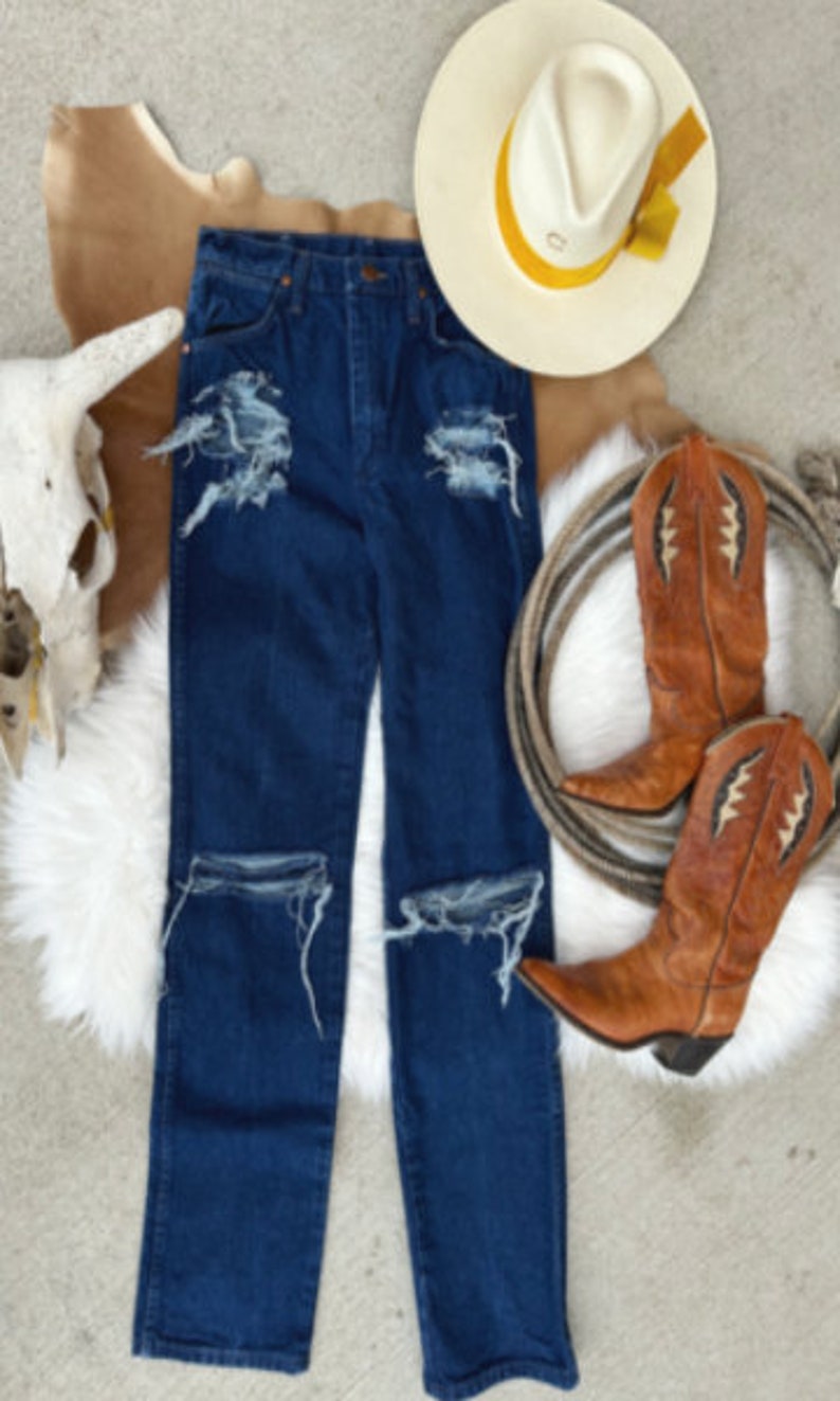 May include: Distressed blue denim jeans with frayed edges and rips at the knees. The jeans are laid flat on a white surface with a cowboy hat and boots in the background.