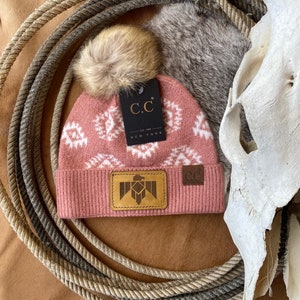 May include: A pink knit beanie with a white geometric pattern and a faux fur pom pom. The beanie has a brown leather patch with a Native American inspired eagle design and the text "C.C. EST. 1998 NEW YORK".