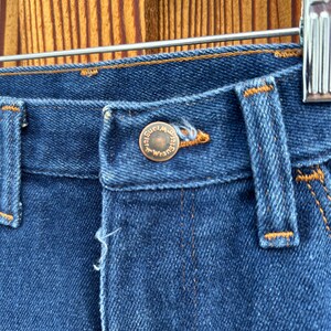 Puede incluir: Primer plano de la parte superior de un par de pantalones vaqueros azules con un bot&oacute;n de color cobre que dice "Wrangler" en un c&iacute;rculo.