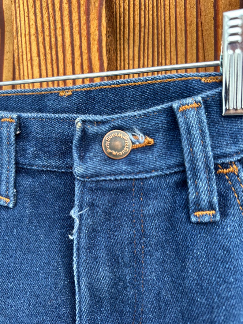 Puede incluir: Primer plano de la parte superior de un par de pantalones vaqueros azules con un bot&oacute;n de color cobre que dice "Wrangler" en un c&iacute;rculo.