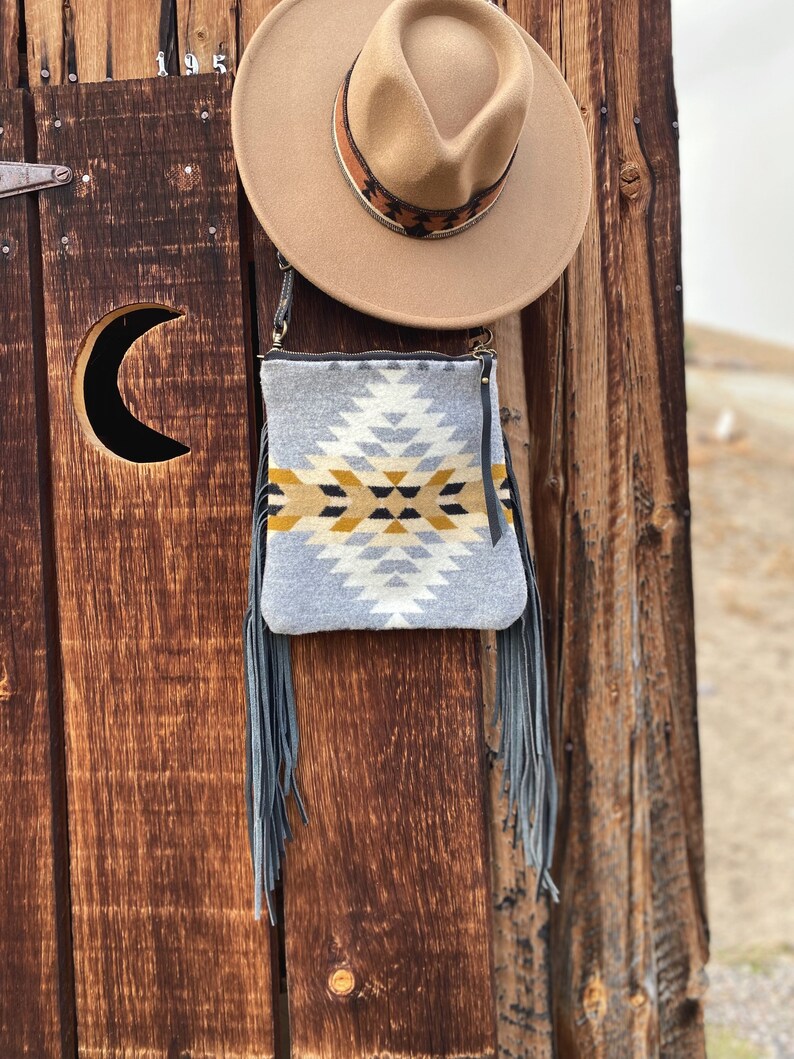 Pode incluir: Uma bolsa crossbody com padr&atilde;o cinza e amarelo com franjas penduradas na parte inferior. A bolsa est&aacute; pendurada em uma porta de madeira com um chap&eacute;u marrom no topo da porta.