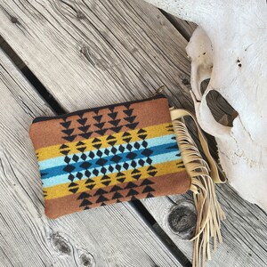 May include: A brown, yellow, blue, and black patterned clutch with a fringe tassel. The clutch is made of a soft, woven fabric and has a zipper closure.