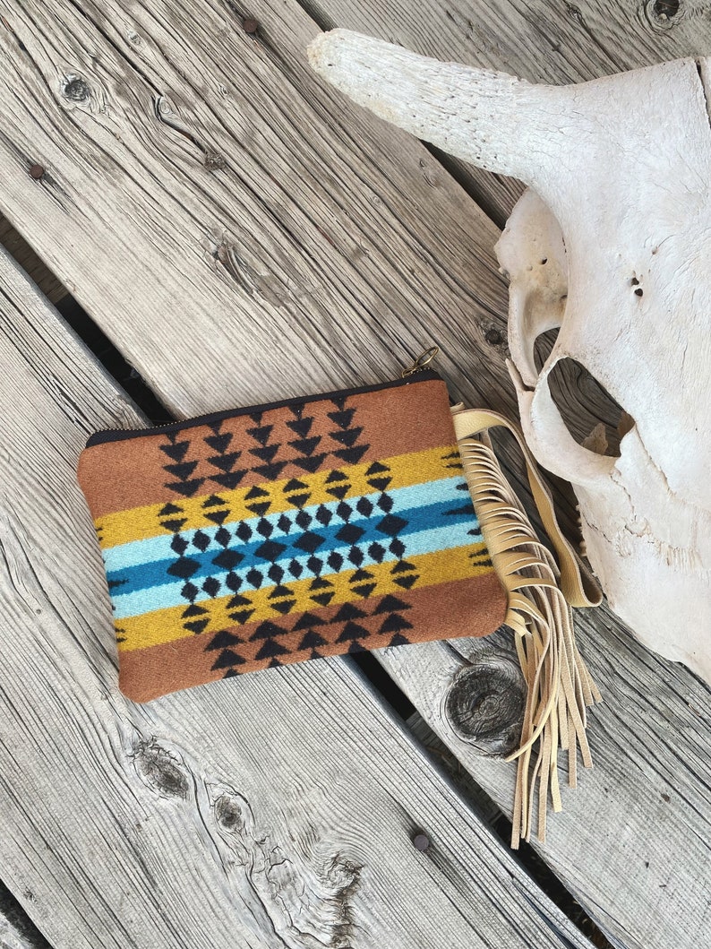 May include: A brown, yellow, blue, and black patterned clutch with a fringe tassel. The clutch is made of a soft, woven fabric and has a zipper closure.