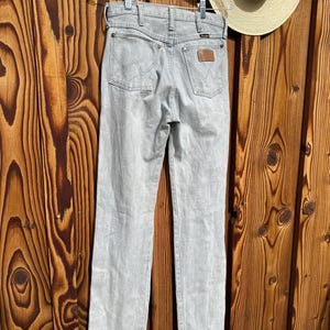 Pu&ograve; includere: Jeans Wrangler grigio chiaro appesi a una gruccia trasparente. I jeans hanno un classico design a cinque tasche con il logo Wrangler sulle tasche posteriori e una toppa in pelle marrone. Un cappello di paglia con una fascia nera &egrave; sullo sfondo.
