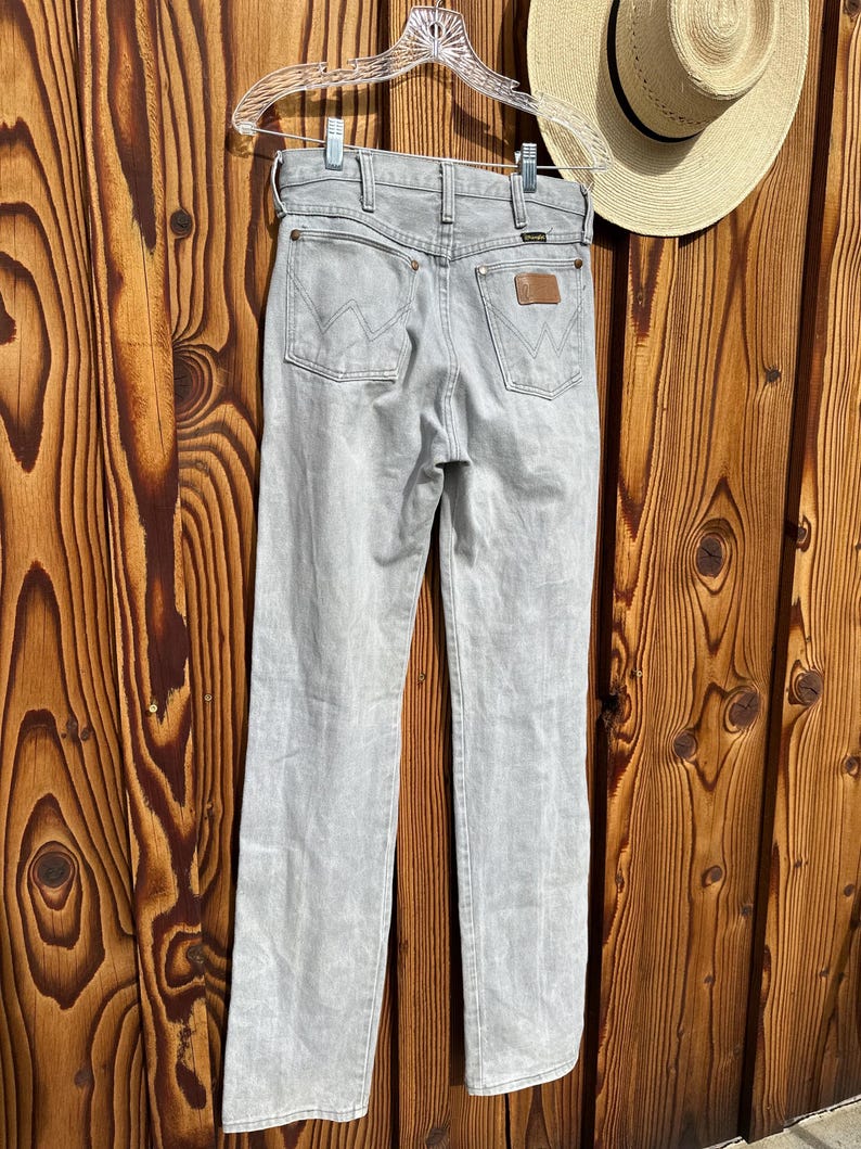 Pu&ograve; includere: Jeans Wrangler grigio chiaro appesi a una gruccia trasparente. I jeans hanno un classico design a cinque tasche con il logo Wrangler sulle tasche posteriori e una toppa in pelle marrone. Un cappello di paglia con una fascia nera &egrave; sullo sfondo.