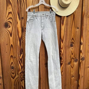 Pu&ograve; includere: Jeans di denim grigio chiaro appesi a una gruccia trasparente su uno sfondo di legno. I jeans hanno un classico design a cinque tasche e una gamba dritta. Un cappello di paglia con una fascia nera &egrave; appeso vicino. I jeans sono lavati chiari.