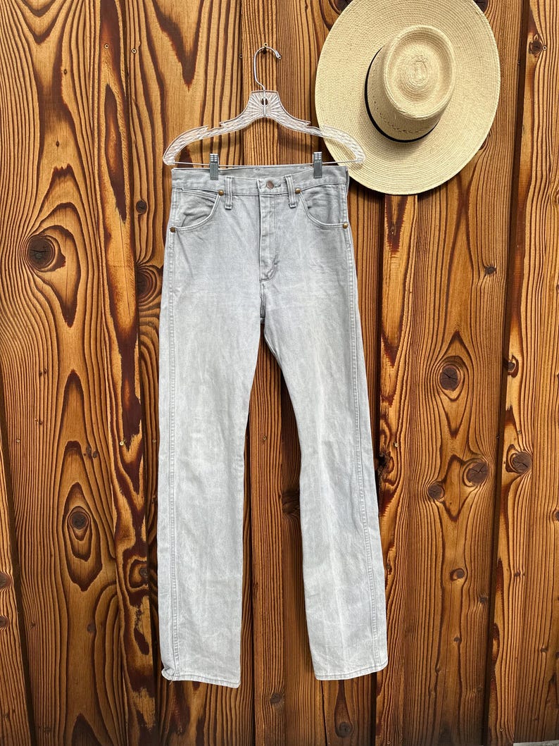 Pu&ograve; includere: Jeans di denim grigio chiaro appesi a una gruccia trasparente su uno sfondo di legno. I jeans hanno un classico design a cinque tasche e una gamba dritta. Un cappello di paglia con una fascia nera &egrave; appeso vicino. I jeans sono lavati chiari.